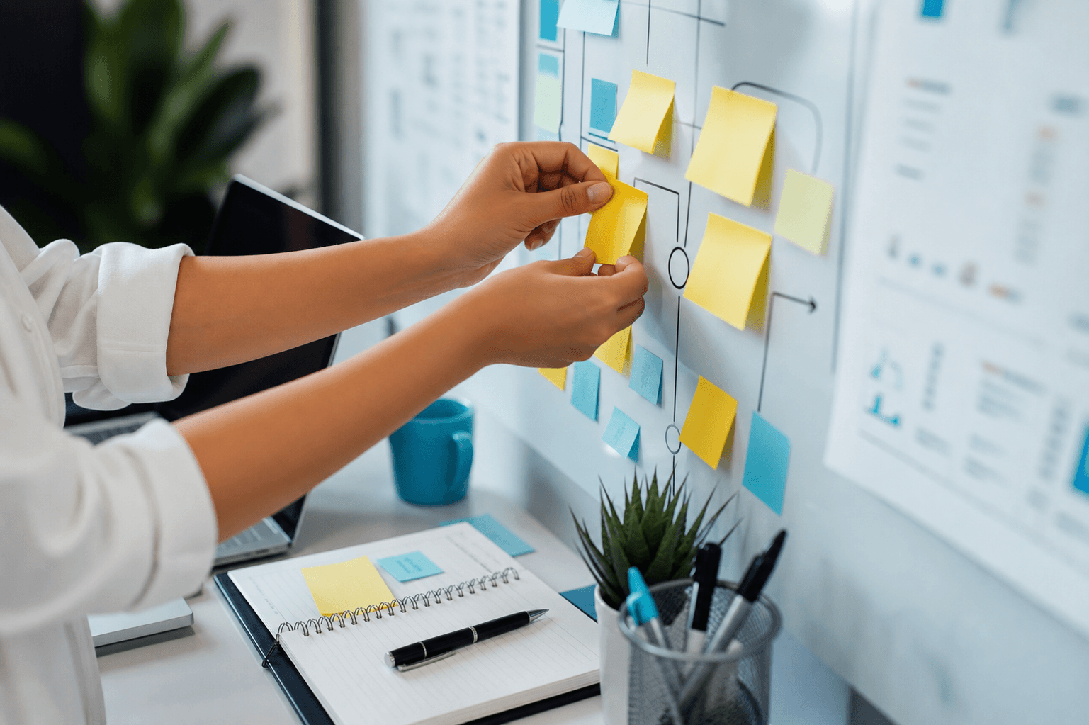 Hands arranging yellow sticky notes on a workflow board beside a notebook and laptop.