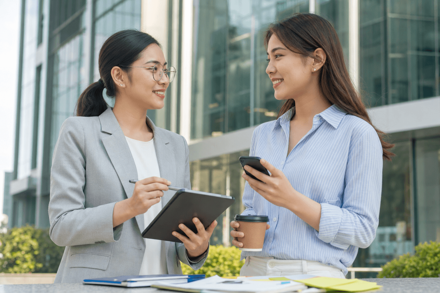 Two businesswomen reviewing social media plans on a phone and tablet outside a modern building.